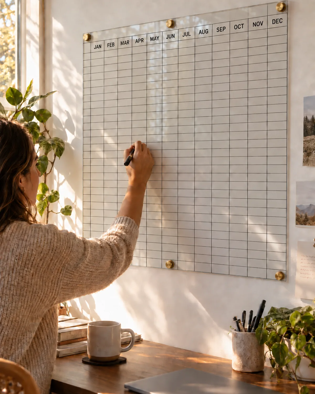 Meter-wide clear acrylic yearly wall planner with a one-color back-printed year grid, standoff-mounted on a plasterboard wall in a home office, with a hand writing in one day cell