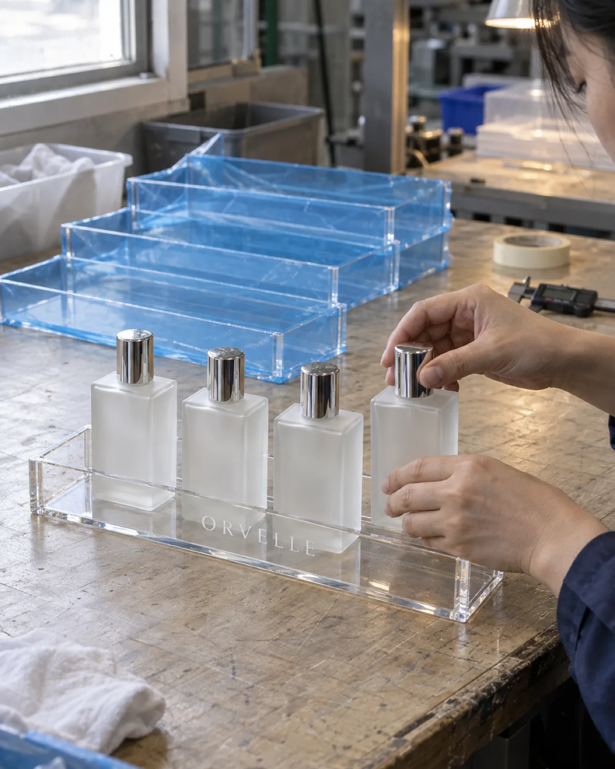 QC inspector seating the fourth frosted-glass bottle into a clear 4 mm acrylic four-bottle perfume tray with a laser-etched wordmark on the front face, more trays in protective film behind