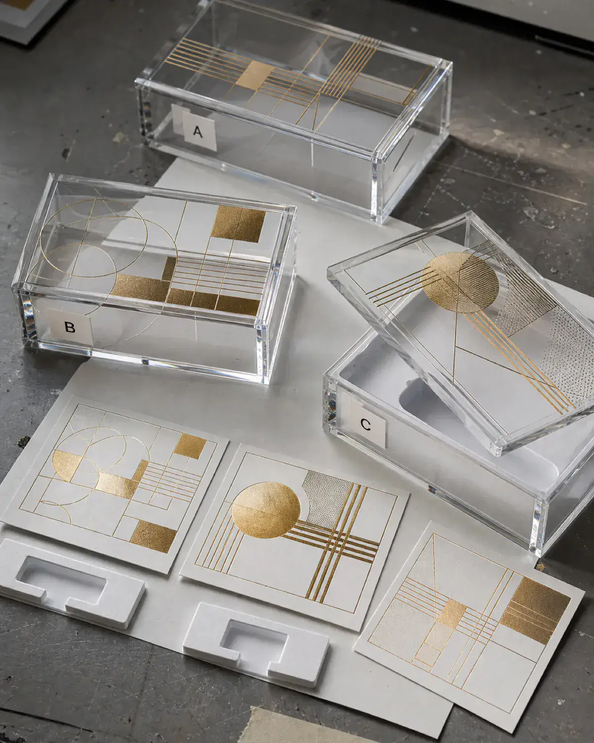 Three clear acrylic presentation boxes with abstract metallic-gold artwork, white insert samples, and A, B, and C version labels on a print bench