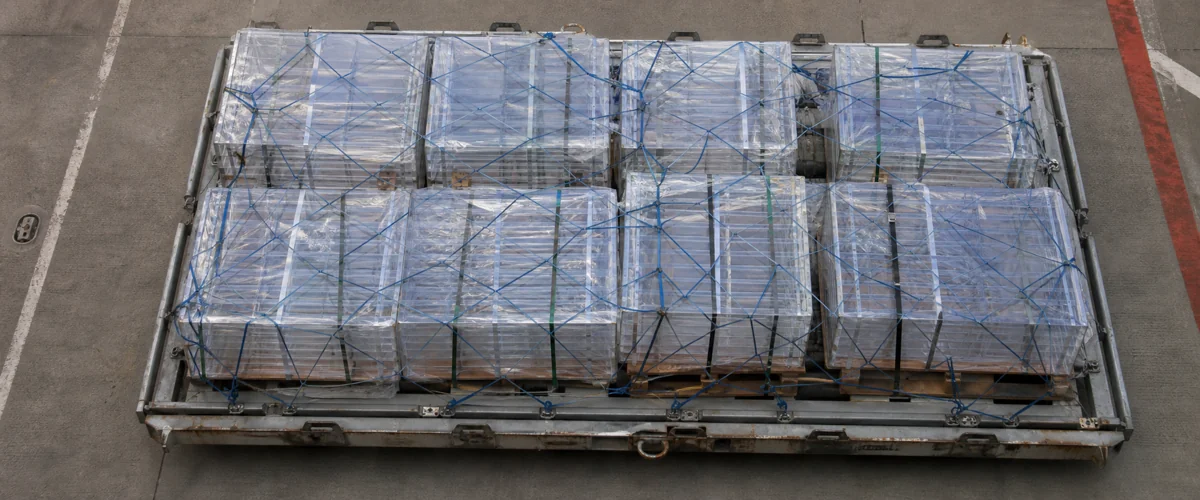 Top-down view of an air-cargo aluminum ULD pallet on the tarmac, loaded with stretch-wrapped acrylic display crates secured under blue netting