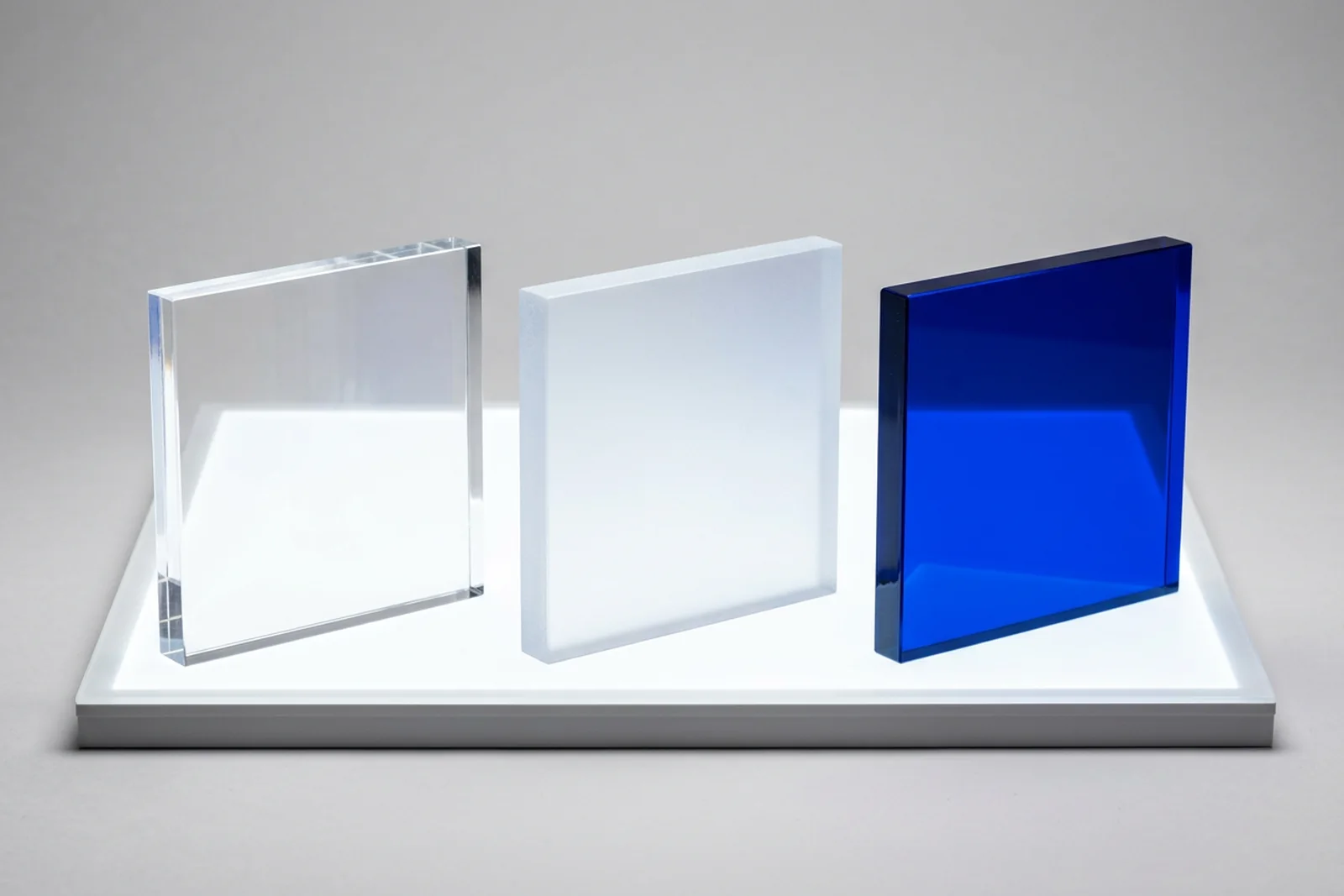 Three acrylic (PMMA plexiglass / perspex) sample squares - clear, frosted, and a deep colored tint - lit from below on a studio light panel, showing the visible transmission difference between finishes.