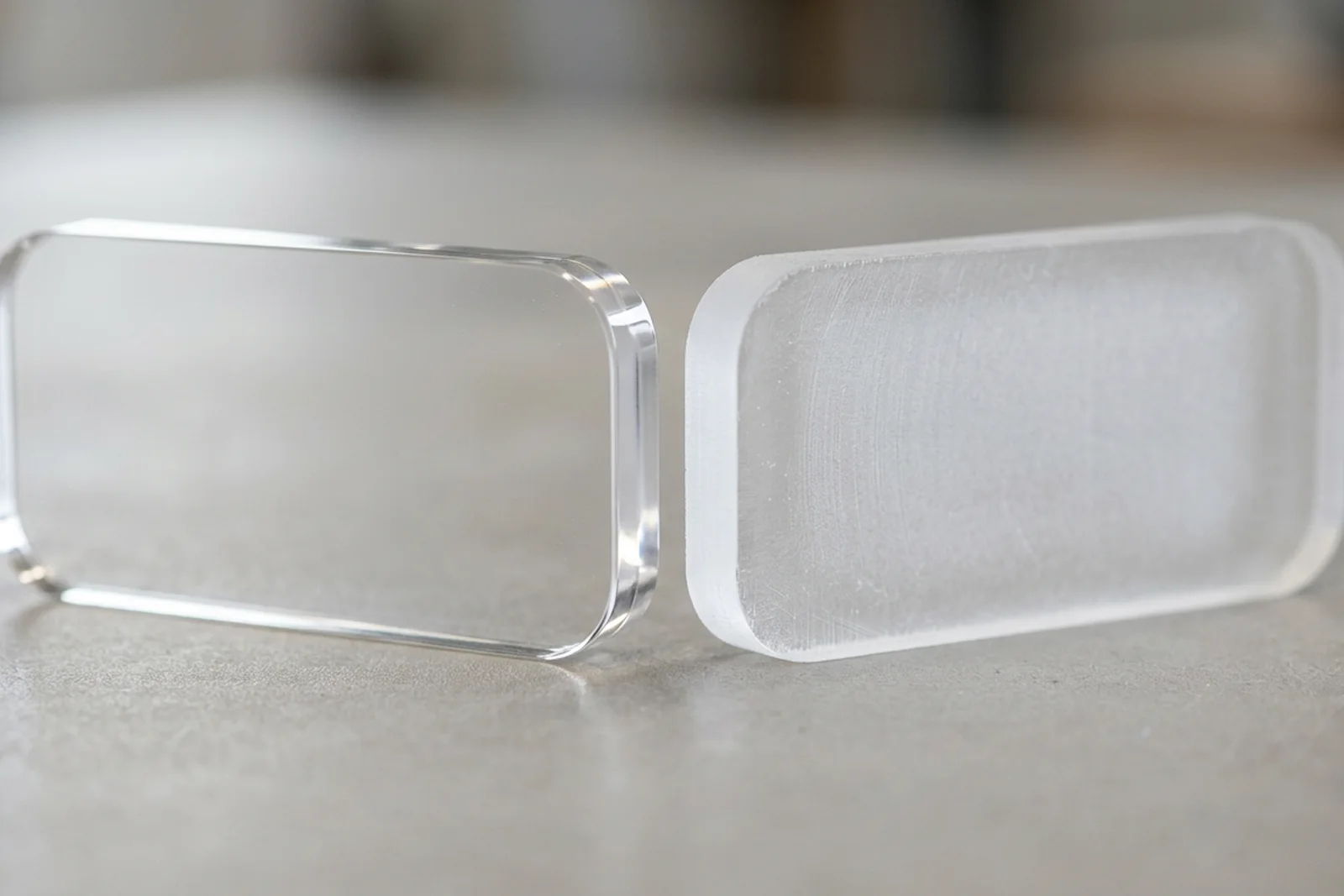 Two small 5mm cast PMMA acrylic (plexiglass) test pieces side by side - the laser-cut piece shows a smooth transparent edge, the CNC-routed piece shows a matte pre-polish edge. Neutral surface, diffused studio light.