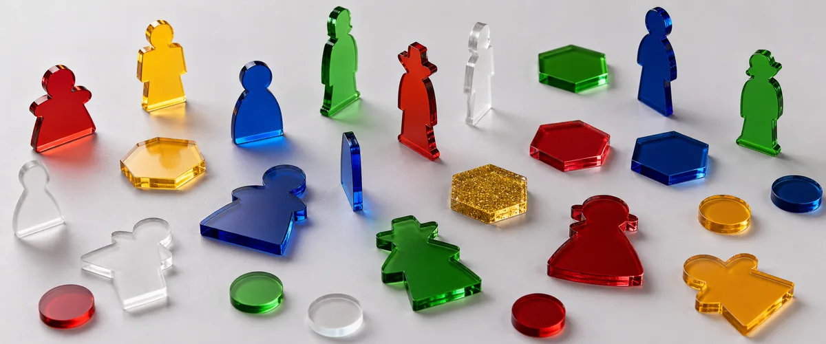 Assorted custom board game pieces in laser-cut colored acrylic — translucent player tokens, engraved markers, and frosted counters arranged on a neutral studio surface