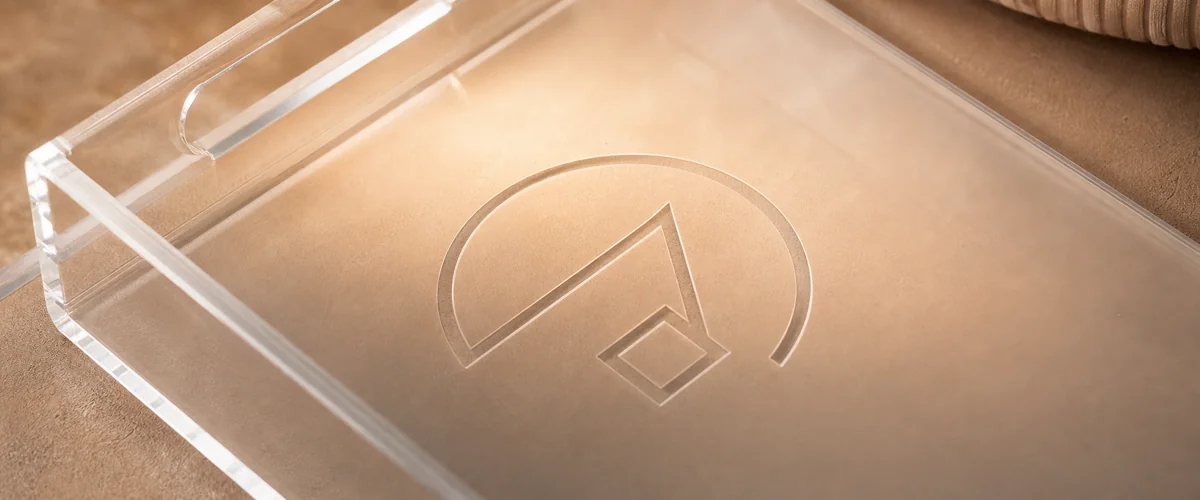 Macro close-up of an etched brand logo on a clear acrylic tray surface, raking light revealing the etched contour and depth