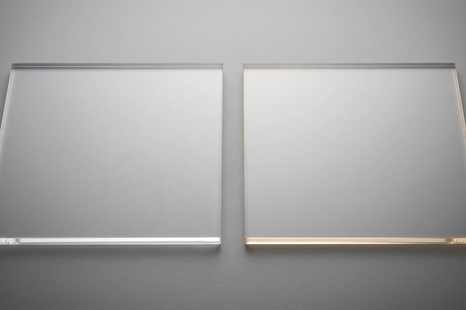 Close-up of cast acrylic (PMMA plexiglass) sample panel next to a polycarbonate (Lexan PC) sample panel on a neutral studio background — acrylic shows crisp edge clarity, polycarbonate shows faint amber tint along the edge
