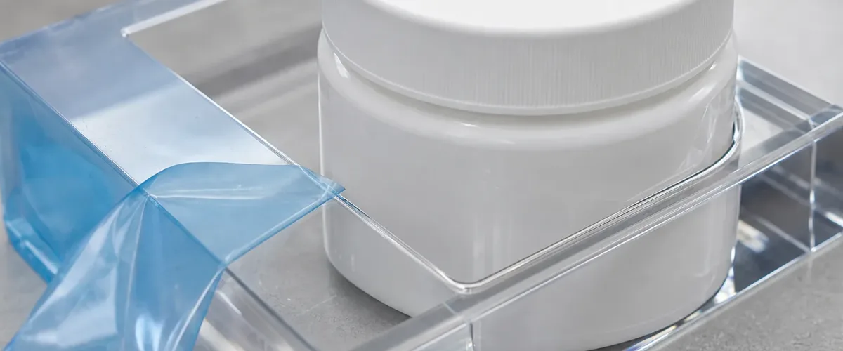 White unlabeled product jar seated in the well of a clear acrylic display sample during a pre-production fit test, blue protective film peeled back from the polished PMMA panel