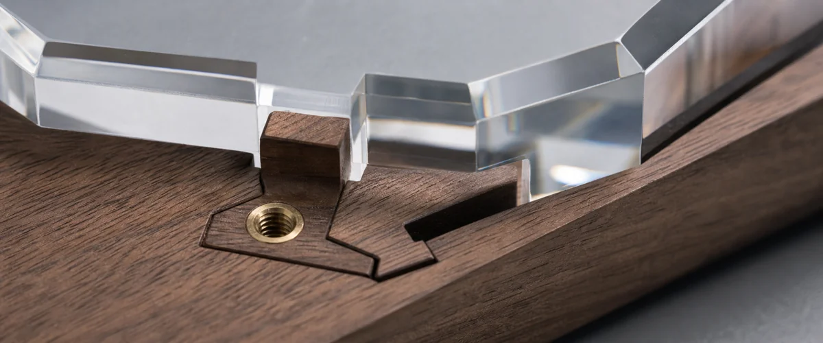 Extreme macro close-up of the joint detail where walnut wood base meets faceted clear acrylic, showing the dovetail interlock and embedded threaded brass insert under directional side light