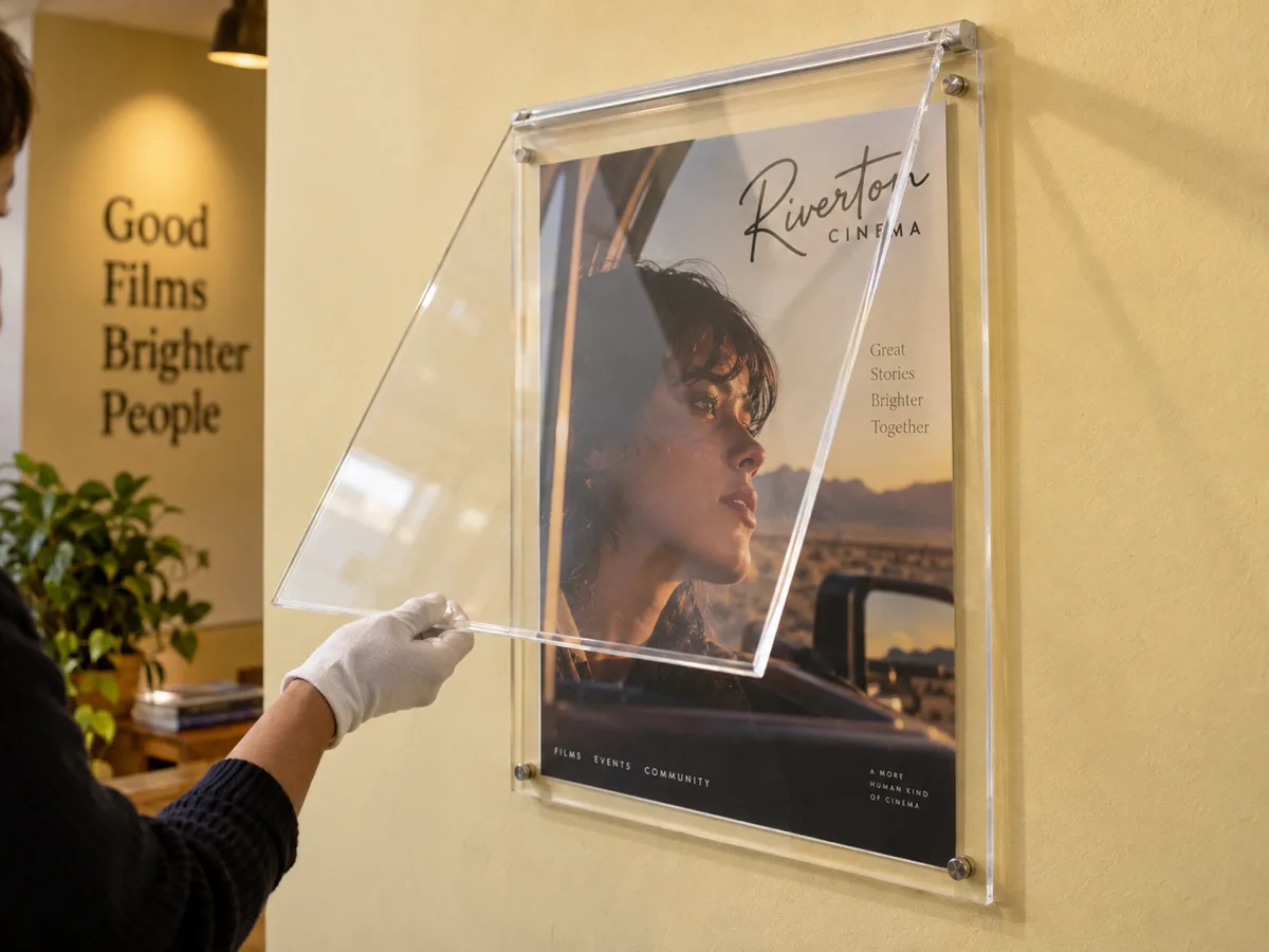 Gloved hand lifting the clear front cover of a wall-mounted acrylic poster case on its top hinge rail